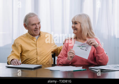 Senior Paar hält Umschlag mit "Roth IRA" Schriftzug und Geld beim Sitzen am Tisch Stockfoto