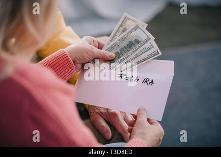 7/8-Ansicht von Senior Paar hält Umschlag mit "Roth IRA" Schriftzug und Dollar Banknoten Stockfoto