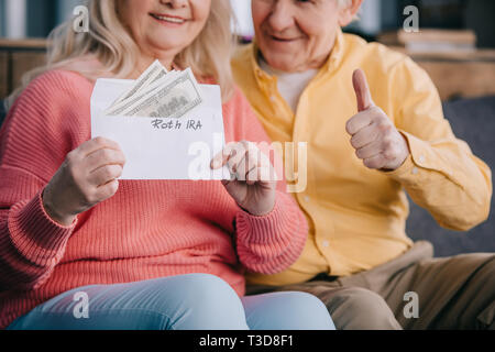 Blick auf ältere Paare mit Daumen hoch, 7/8-Zeichen bei gedrückt gehaltener Umschlag mit "Roth IRA" Schriftzug und Dollar Banknoten Stockfoto