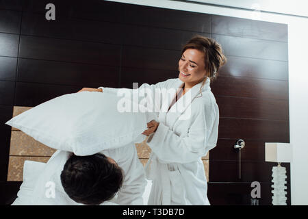 Glückliches Paar in Bademänteln in Kissenschlacht im Bett im Hotel Stockfoto