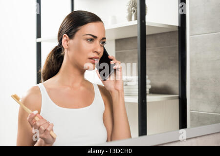 Selektiver Fokus der schöne Brünette Frau mit Zahnbürste während des Sprechens auf dem Smartphone im Badezimmer Stockfoto
