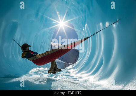 Tief in den alaskischen Wildnis, ein Ice climber ruht in einer Hängematte in die Wände von einer Eishöhle. Die Sonne scheint in den Eingang der Großen Blauen c Stockfoto