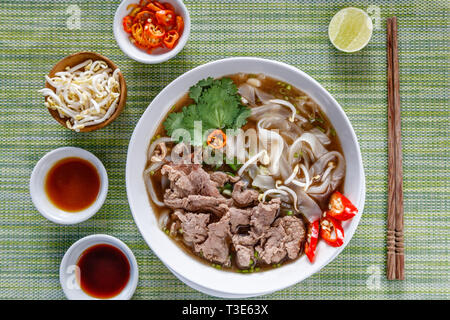 Vietnamesischen traditionellen Rindfleisch und Nudelsuppe Pho Bo in Weiße, runde Schüssel serviert, mit Saucen, Kalk, Chili und Sojasprossen. Holz- Stäbchen. Stockfoto