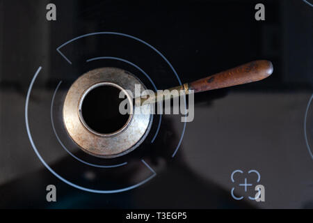 Ansicht von oben alte Bronze Türkische retro Kaffeemaschine kanaka auf Glas Herd und Backofen mit Griff aus Holz, auf einem schwarzen Hintergrund in die Küche getragen. Retro cezv Stockfoto