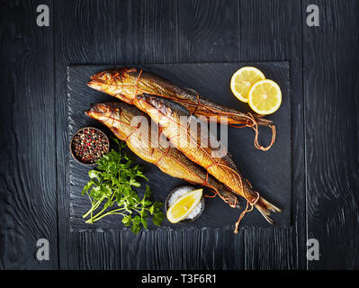 Drei heiß geräucherten Heringe auf schwarzem Schiefer Fach mit Zitronenscheiben, Petersilie und Pfefferkörner, Ansicht von oben, flatlay Stockfoto