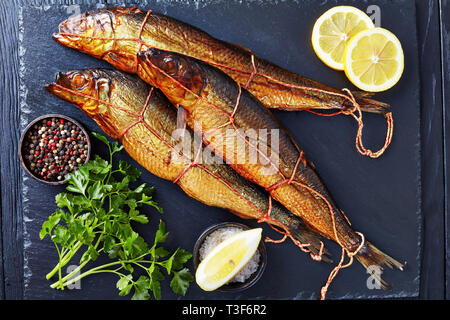 Drei heiß geräucherten Heringe auf schwarzem Schiefer Fach mit Zitronenscheiben, Petersilie und Pfefferkörner, Ansicht von oben, flatlay, close-up Stockfoto