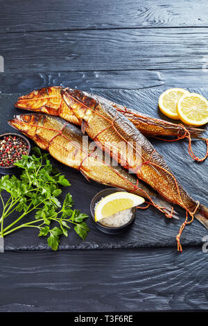 Heiß geräucherte ganze Heringe mit Zitronenscheiben, Petersilie und Pfeffer auf einem schwarzen Schiefer Fach auf einem Holztisch, vertikale Ansicht von oben, Kopie Raum Stockfoto