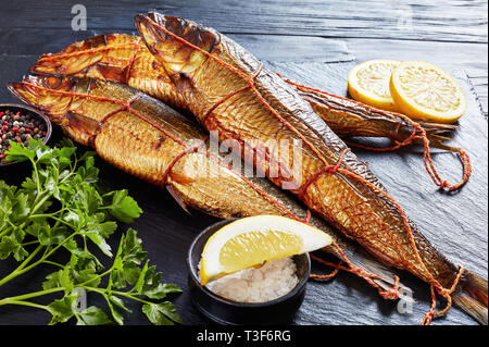 Frisch geräucherte ganze Heringe mit Zitronenscheiben, Petersilie und Pfeffer auf einem schwarzen Schiefer Fach auf einem Holztisch, horizontale Ansicht von oben Stockfoto