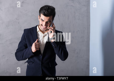 Ein glücklicher hübscher junger Geschäftsmann in einem schwarzen Anzug über sein Telefon vor einer grauen Wand in einem Studio. Stockfoto