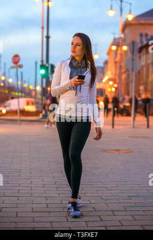 Eine schöne junge Frau tragen Sportswear und stehen mit ihrem Handy auf der Straße in der Stadt-Lichter in der Nacht fällt Stockfoto