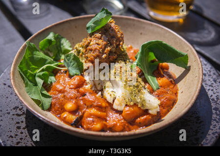Ein warmes weißes Fischfilet auf Bohnen in Tomatensauce mit grünen Blättern serviert. Stockfoto