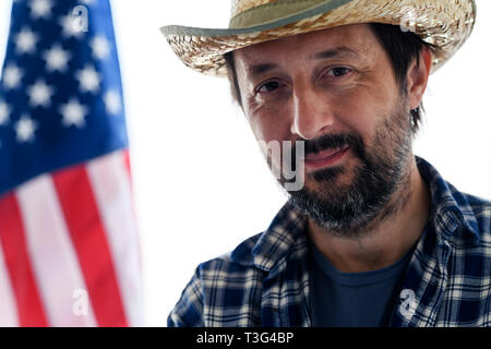 Lächelnd amerikanischen Landwirt mit Kamera suchen, Portrait von erwachsenen männlichen Person mit Strohhut und Plaid Shirt mit USA-Flagge im Hintergrund Stockfoto