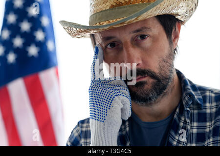 Nachdenklich amerikanischen Landwirt mit dem Finger auf die Stirn an Kamera suchen, Portrait von erwachsenen männlichen Person mit Strohhut und Plaid Shirt mit USA-Flagge in Ba Stockfoto