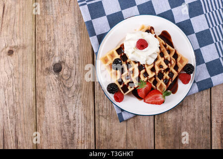 Frische belgische Waffeln mit Schokoladensauce, Erdbeeren, Obst und Beeren und Sahne auf rustikalen Holztisch Stockfoto