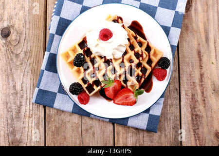 Frische belgische Waffeln mit Schokoladensauce, Erdbeeren, Obst und Beeren und Sahne auf rustikalen Holztisch Stockfoto