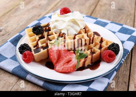 Frische belgische Waffeln mit Schokoladensauce, Erdbeeren, Obst und Beeren und Sahne auf rustikalen Holztisch Stockfoto