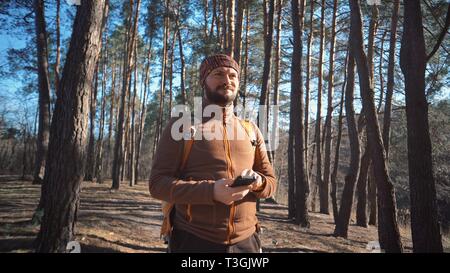 Thema Tourismus und Technologie. Jungen kaukasischen Mann mit Bart und Rucksack. Wandern touristische in einem Pinienwald verwendet die Technologie, Hand Handy Stockfoto