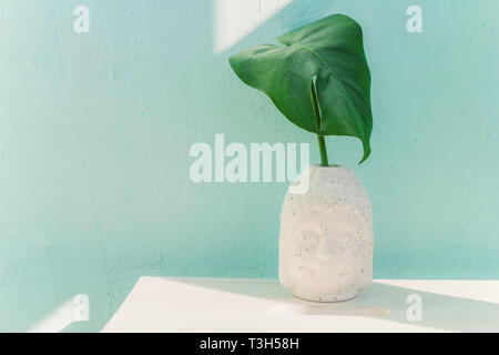 Grüne monstera Blatt in Keramik Buddha Kopf Vase auf blauen Wand Hintergrund, Sommer tropische Minimalismus Kopie Raum Stockfoto