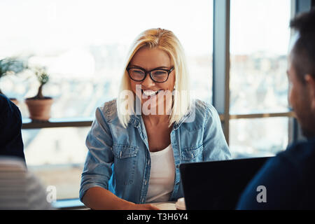 Junge blonde Frau lachend während der Sitzung gegenüber Kollegen an einem Tisch während einer Sitzung in einem modernen Büro Stockfoto