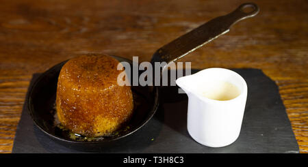 Sticky Toffee Pudding serviert mit Karamellsauce und ein Krug mit Vanillepudding. Das Dessert wird in der Pfanne serviert. Stockfoto