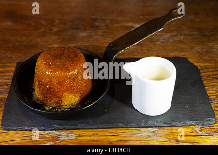 Sticky Toffee Pudding serviert mit Karamellsauce und ein Krug mit Vanillepudding. Das Dessert wird in der Pfanne serviert. Stockfoto