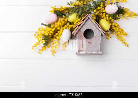 Vogel Haus Frühling Blumen Mimosa und dekorative Ostereier auf weißem Holz- Hintergrund Stockfoto