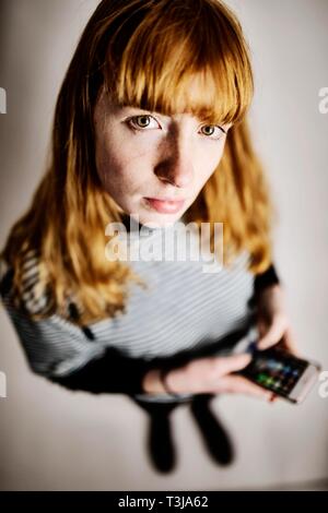 Mädchen, Teenager, Rothaarige, mit Smartphone in der Hand, schaut skeptisch in Kamera, studio Shot, Deutschland Stockfoto