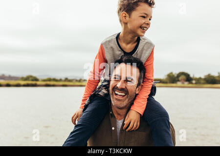 Kind sitzen auf den Schultern seines Vaters und Spaß haben. Porträt eines lächelnden Mann mit seinem Kind sitzen auf den Schultern in der Nähe von einem See. Stockfoto