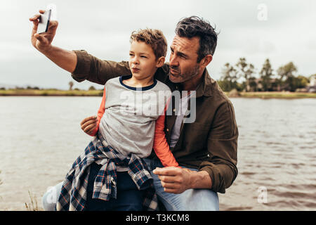 Nahaufnahme von Vater und Sohn eine selfie sitzen in der Nähe von einem See. Glückliches Kind sitzt auf dem Schoß seines Vaters Blick auf das Mobiltelefon, während seine fathe