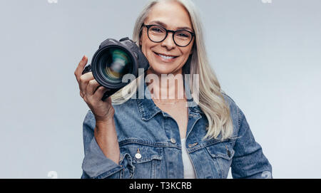 Zuversichtlich ältere Frau in casuals Holding eine DSLR-Kamera auf grauem Hintergrund. Professionelle Fotografin mit Digitalkamera. Stockfoto