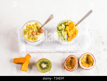 Zwei Gläser mit weißen Joghurt und Obst Pürieren von Passionsfrucht, Kiwi, Mango und mit metallischen Löffel und halbe Früchte auf weiße strukturierte Hintergrund. Top Stockfoto