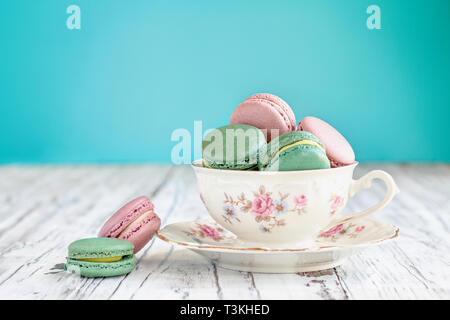 Antike Bavaria Winterling footed Tasse Tee aus den 1950er Jahren "gefüllt mit rosa Erdbeere und grüner Tee macarons auf einem urigen weißer Tisch gegen einen Teal ba Stockfoto