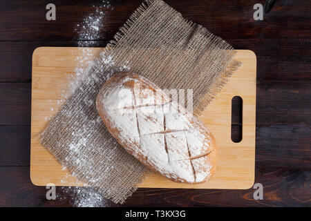 Frisches Brot auf einer Eiche board mit verstreuten Mehl auf einer hölzernen Hintergrund mit einem Stück Leinen Stoff. Stockfoto