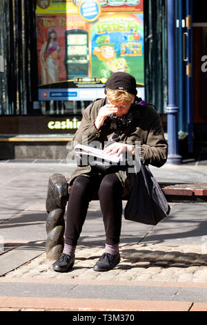 Eine ältere Frau sitzt auf einem Sitz in der Sonne tun die Zeitung Kreuzworträtsel in Dundee, Großbritannien Stockfoto