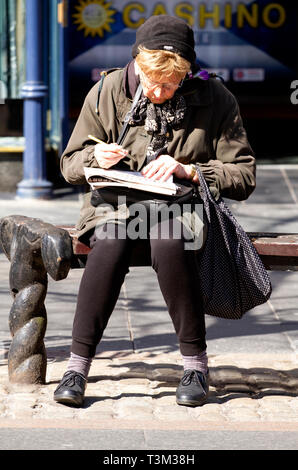 Eine ältere Frau sitzt auf einem Sitz in der Sonne tun die Zeitung Kreuzworträtsel in Dundee, Großbritannien Stockfoto