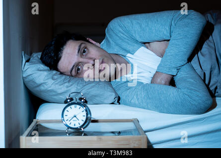 Schlaflose und verzweifelten jungen kaukasischen Mann wach nachts nicht schlafen zu können, frustriert und beunruhigt, schaut auf die Uhr leiden unter Schlaflosigkeit. Stockfoto