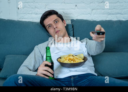 Lifestyle Portrait der junge Mann gelangweilt auf der Couch mit Fernbedienung zapping für Film oder Sport live. Suchen desinteressiert Bier trinkt. Bewegungsmangel und Stockfoto