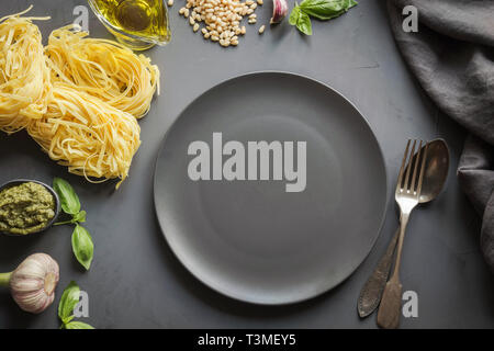 Leere schwarze Platte und Grenze der rohen Pasta, Pesto, Basilikum, Parmesan zum Kochen mediterrane Gerichte auf dunklen konkreten Hintergrund. Ansicht von oben, Platz für de Stockfoto