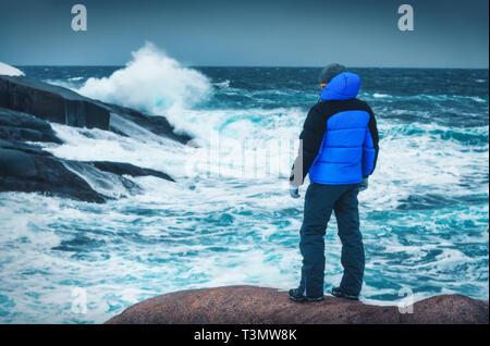 Sportlicher Mann in der blauen Jacke steht auf dem Stein an der Küste mit Felsen und grosse Wellen im Winter. Kalte bewölkten Tag in Teriberka, Russland. Dramat Stockfoto