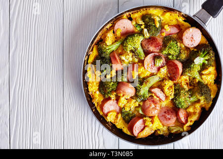 Leckere heiße Omelette mit Brokkoli und Würste in einer Pfanne auf einem weißen Holztisch, horizontale Ansicht von oben, flach, Nahaufnahme, Kopie Raum Stockfoto