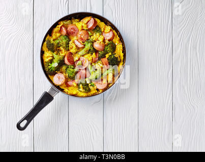 Leckere heiße Omelette mit Brokkoli und Würste in einer Pfanne auf einem weißen Holztisch, horizontale Ansicht von oben, flach, leeren Raum rechts Stockfoto