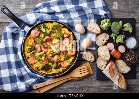 Leckere heiße Omelette mit Brokkoli und Würste in einer Pfanne auf einem alten Holztisch mit Zutaten in Scheiben geschnitten und knusprigem Baguette Vollkorn, Stockfoto