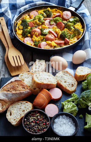 Köstliche warme Rühreier mit Brokkoli und Würste in einer Pfanne auf einem alten Holztisch mit Zutaten in Scheiben geschnitten und crusty Vollkorn Französische bagu Stockfoto