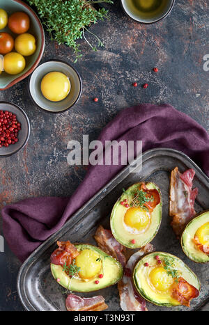 Avocado mit Ei und Speck auf einem Backblech gebacken, flach mit Zutaten und Kräuter auf dunklem Hintergrund Stockfoto