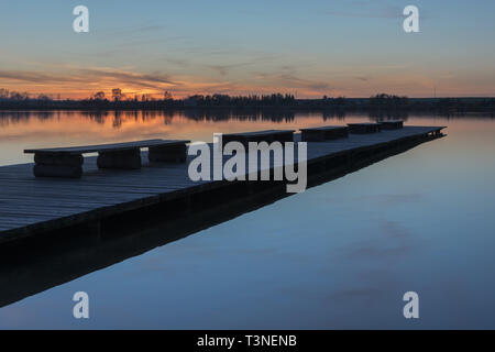 Eine lange Brücke mit Bänken, Sonnenuntergang über einem ruhigen See Stockfoto