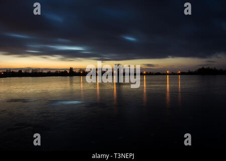 Dunkle Wolken nach Sonnenuntergang auf einem zugefrorenen See und reflektierende Lichter im Eis - Twilight anzeigen Stockfoto