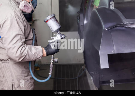 Ein Mann in Schutz Overalls und eine Maske hält eine Sprayflasche in der Hand und spritzt schwarze Farbe auf den Rahmen der Karosserie nach einem Unfall durin Stockfoto