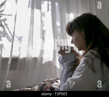 Junge Frau trinkt einen Kaffee, während Sie im Bett sitzen, gegen das Fenster Stockfoto
