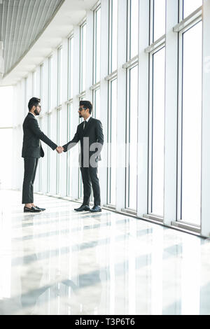 Zwei indische Unternehmer die Hände schütteln bei grossen Fenster mit urbanen Stadtlandschaft, zuversichtlich Geschäftspartner handshaking im Amt. Seitenansicht Stockfoto