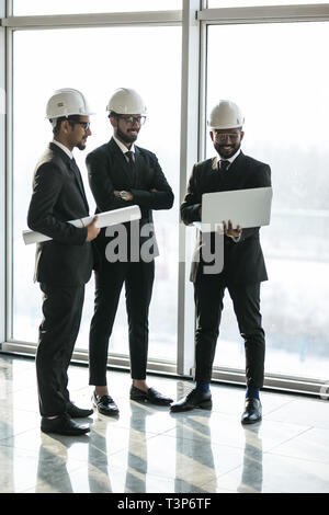 Nahaufnahme von drei lächelnd diverse Geschäftsleute Helme tragen und stehen im Freien mit Gebäude im Hintergrund. Architekten Konzept. Stockfoto
